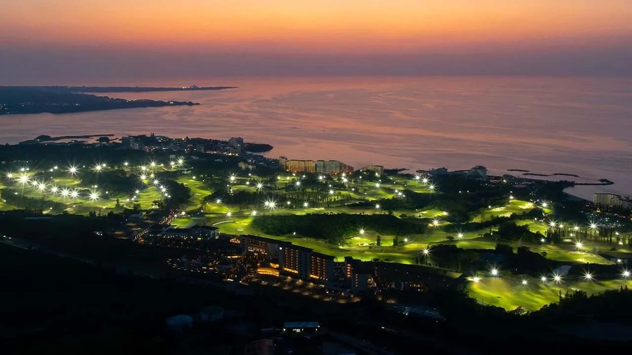 マジックアワーの空と海に包まれる、PGMホテルリゾート沖縄のドラマチックな夕景。ライトアップされた広大なゴルフコースが宝石のように輝く、心奪われる絶景リゾート